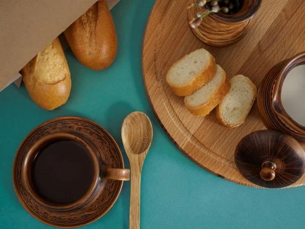 Breakfast in handmade earthenware top view. Cup of tea, milk in a jug, baguette in a paper bag, on a wooden tray pussy willow branches in a vase, wooden spoon on a green background. Flat lay