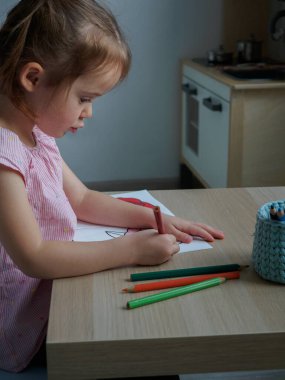 4 yaşındaki küçük kız odasındaki ahşap bir masaya boyama kitabını özenle çiziyor.