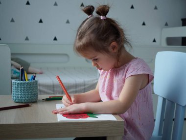 Pembe çizgili tişörtlü güzel bir kız evindeki odasındaki ahşap masa desenine göre kalemle bir gül boyuyor.