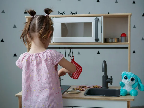 4-5-letnia dziewczynka w różowej koszulce w paski bawi się w swoim pokoju w kuchni dla dzieci z patelniami na zabawki — Zdjęcie stockowe