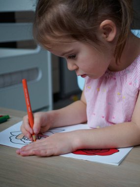 4 yaşındaki çekici bir kız, evindeki odasındaki ahşap bir masada turuncu kalemle bir çiçek resmi çiziyor.
