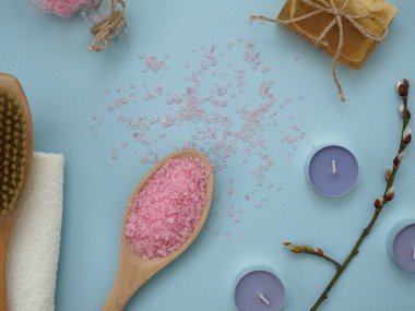 Mavi arka planda spa tedavileri için aksesuarlar. Tahta kaşıkta deniz tuzu, kuru masaj fırçası, el yapımı sabun, beyaz havlu, mavi renkli çay mumları, üst manzara, düz yatak.
