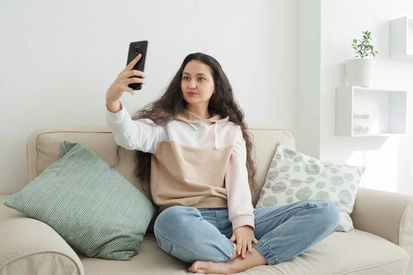 Asyalı görünümlü genç bir kadın blogu için evdeki kanepede selfie çekiyor.