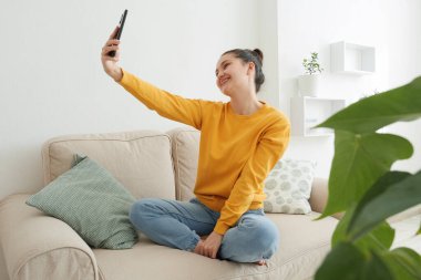 Genç ve güzel bir kadın dairedeki koltukta otururken telefonda selfie çekiyor.