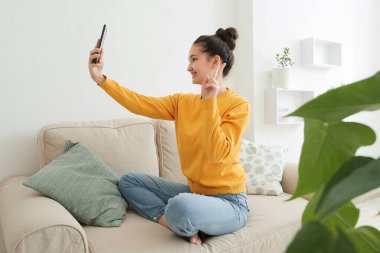 Sarı kazaklı Asyalı kız evde koltukta selfie çekiyor.