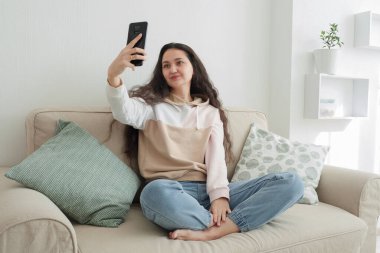Uzun kıvırcık saçlı güzel Asyalı kız elinde telefonla kanepede oturur ve selfie çeker.