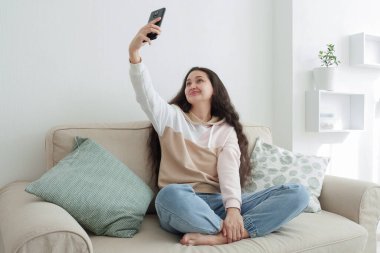Asya görünümlü uzun saçlı genç bir kadın evdeki kanepede selfie çekiyor. İnternette hayat.