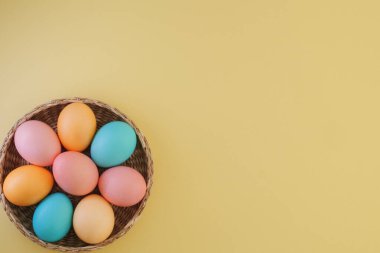 Paskalya minimalist arka planı, sarı arka planda sepette pastel renkli Paskalya yumurtaları. Boşluğu kopyala