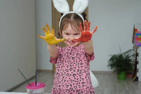 4-5 yaşlarında pembe elbiseli ve beyaz Paskalya tavşanı kulaklı bir kız suluboya ellerle dolu bir odada dikiliyor.