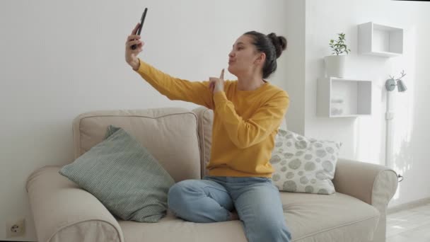 En charmerende kvinde blogger kommunikerer med sine abonnenter på en smartphone live. Asiatisk kvinde i gul sweatshirt og jeans sidder på sofaen derhjemme – Stock-video