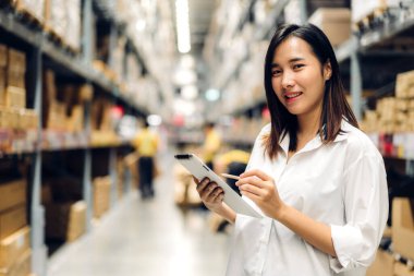 Asyalı yönetici kadın portresi ayakta duruyor ve malzeme ve gereçleri depoda kontrol etmek için tablet bilgisayardan ayrıntılı talimat veriyor. Lojistik ve iş ihracatı