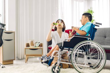 Mutlu aşk portresi. Asyalı anne oynuyor ve bakıcı tekerlekli sandalyede oturan engelli çocuğa bakıyor. Evde engelli bakımı konseptinde iyi vakit geçiriyor.