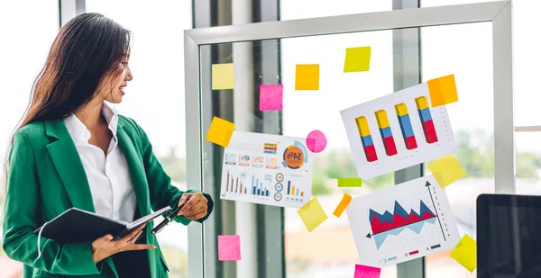 Casual creative happy smile asian business woman planning strategy analysis putting post it stickers note to share idea for startup project on glass board at modern office