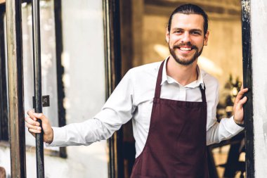 Kahve dükkanının önünde gülümseyen yakışıklı sakallı baristanın portresi. Kafenin önünde duran erkek barmen.