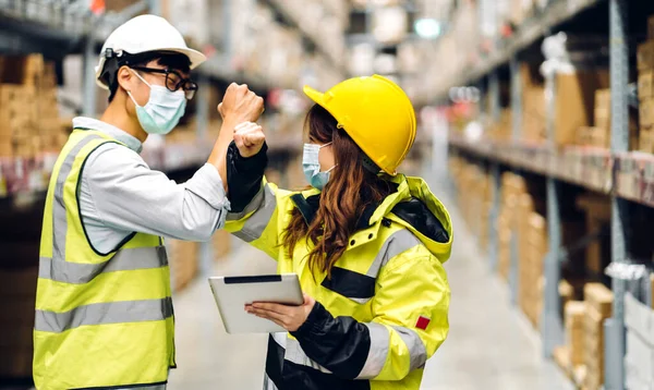 Asian engineer man and woman in helmets in quarantine for coronavirus wearing protective mask shaking with elbows in new normal at shelves with goods background in warehouse.logistic and business export