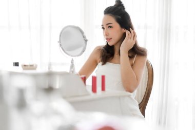 Genç, güzel Asyalı bir kadının, temiz, sağlıklı, beyaz tenli, aynaya bakan, elleriyle yüzüne dokunan ve evde krem süren bir kızın gülümsemesi. Spa ve güzellik konsepti.