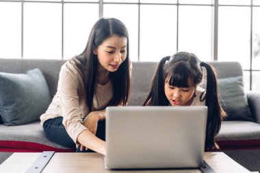 Anne ve Asyalı küçük kız öğreniyor ve dizüstü bilgisayara bakıyor ev ödevi yapıyor çevrimiçi eğitim e-öğrenme sistemiyle bilgi öğreniyor. Evde öğretmenle çocuk video konferansı