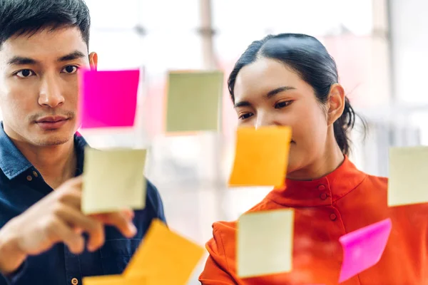 Casual creative happy two smile asian business people planning strategy analysis and brainstorm putting post it stickers note to share idea for startup project on glass board at modern office