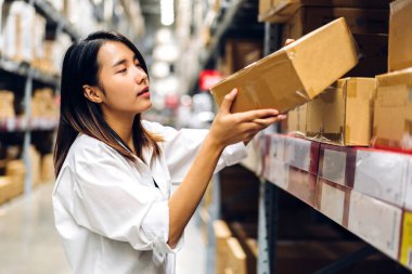 Gülümseyen Asyalı yönetici kadın portresi ayakta duruyor ve ambardaki malzeme ve malzemeleri kontrol etmek için karton kutunun üzerinde ayrıntılı talimatlar veriyor. Lojistik ve iş ihracatı