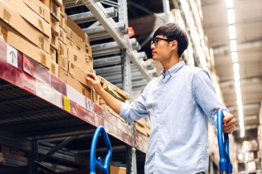 Gülümseyen Asyalı yöneticinin portresi ayakta duruyor ve ambarda karton kutu ürünleri olan raflardaki malzeme ve malzemeleri kontrol ediyor. Lojistik ve ticari ihracat