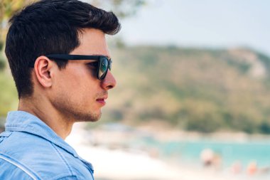 Gülümseyen yakışıklı erkek modelinin portresi, şık hippi yaz gözlükleriyle tropik sahilde durup deniz manzarası izlerken keyif çatmak ve rahatlamak.