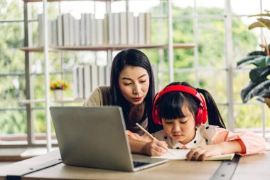 Anne ve Asyalı küçük kız öğreniyor ve dizüstü bilgisayara bakıyor ev ödevi yapıyor çevrimiçi eğitim e-öğrenme sistemiyle bilgi öğreniyor. Evde öğretmenle çocuk video konferansı
