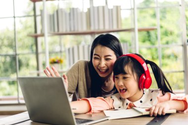 Anne ve Asyalı küçük kız dizüstü bilgisayarda öğreniyor ev ödevi yapıyor çevrimiçi eğitim e-öğrenim sistemiyle. Çocuklar video konferansında öğretmenle evde görüşüyorlar.