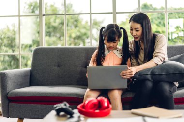 Anne ve Asyalı küçük kız öğreniyor ve dizüstü bilgisayara bakıyor ev ödevi yapıyor çevrimiçi eğitim e-öğrenme sistemiyle bilgi öğreniyor. Evde öğretmenle çocuk video konferansı