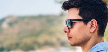 Gülümseyen yakışıklı erkek modelinin portresi. Moda hipster yaz gözlükleriyle keyif alıyor ve rahatlıyor. Tropikal plajlara metin eklemek ve deniz manzarası izlemek için fotokopi çekiliyor.