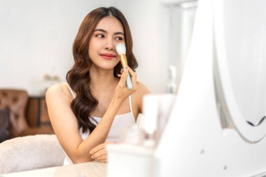 Güzel, güzel Asyalı bir kadının, temiz, sağlıklı, beyaz bir tenin aynaya bakışıyla gülümsemesi. Elinde makyaj fırçası tutan Asyalı bir kız ve yüzünde makyaj malzemesi var. Yüz güzelliği ve kozmetik konsepti.