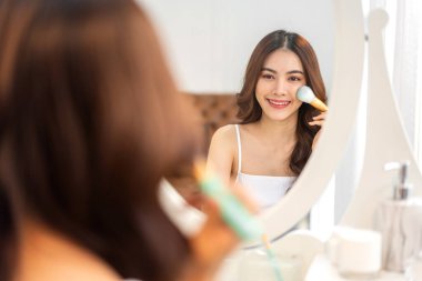 Güzel, güzel Asyalı bir kadının, temiz, sağlıklı, beyaz bir tenin aynaya bakışıyla gülümsemesi. Elinde makyaj fırçası tutan Asyalı bir kız ve yüzünde makyaj malzemesi var. Yüz güzelliği ve kozmetik konsepti.