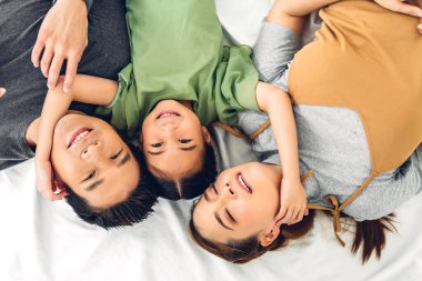 Portrenin en üst görüntüsü mutlu bir şekilde gülümseyen Asyalı bir aile babası ve annesi ve genç bir ebeveyni olan Asyalı bir kız çocuğu dakikalar içinde kameraya bakıyor ve evde yerde uzanıyor.