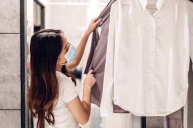 Koronavirüs için karantinaya alınan genç ve güzel Asyalı bir kadın, cerrahi maske takarak korunuyor. Sosyal uzaklık alışverişi yapıyor ve mağazadan kıyafet seçiyor. Covid19 ve yeni normal kavram