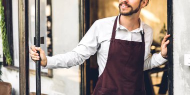 Kahve dükkanının önünde gülümseyen yakışıklı sakallı baristanın portresi. Kafenin önünde duran erkek barmen.