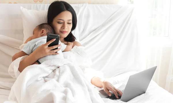 Portrait of enjoy love family asian mother relaxing using technology of laptop computer with asian baby son while sitting on bed.Young creative girl working at home.work from home concept