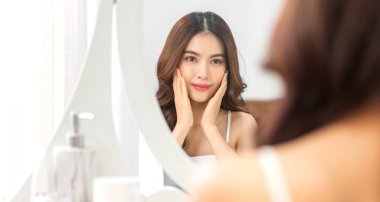 Genç, güzel Asyalı bir kadının, temiz, sağlıklı, beyaz bir tenle aynaya bakması. Asyalı bir kızın elleriyle yüzüne dokunması ve evde krem sürmesi. Spa ve güzellik konsepti.