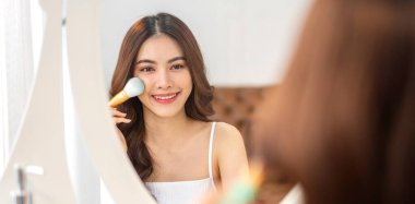 Güzel, güzel Asyalı bir kadının, temiz, sağlıklı, beyaz bir tenin aynaya bakışıyla gülümsemesi. Elinde makyaj fırçası tutan Asyalı bir kız ve yüzünde makyaj malzemesi var. Yüz güzelliği ve kozmetik konsepti.