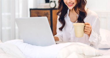 Genç gülümseyen mutlu Asyalı kadın dizüstü bilgisayar kullanarak rahatlıyor ve evdeki yatak odasında kahve içiyor. Genç yaratıcı kız klavyede çalışıyor ve daktilo kullanıyor. Ev konseptinden çalışıyor.