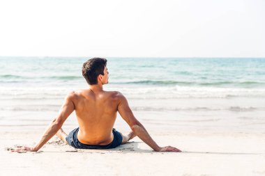 Gülümseyen yakışıklı seksi adamın portresi kaslı fit vücudunu gösteriyor tropik sahilde durup keyif çatıyor ve deniz manzarası seyrediyor.