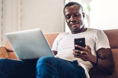 Genç siyahi Afrikalı adam dizüstü bilgisayarla evde çalışıyor ve video konferansı yapıyor. Genç yaratıcı Afrikalı adam akıllı telefonla konuşuyor. Ev konseptinden çalışıyor.