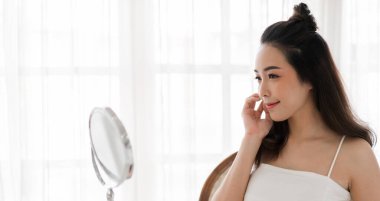 Genç, güzel Asyalı bir kadının, temiz, sağlıklı, beyaz bir tenle aynaya bakması. Asyalı bir kızın elleriyle yüzüne dokunması ve evde krem sürmesi. Spa ve güzellik konsepti.