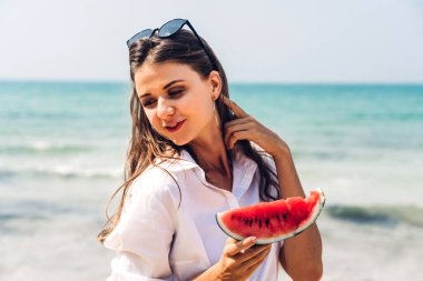 Gülümseyen bir kadının portresi tropik sahilde kucağında bir dilim karpuz yiyor.