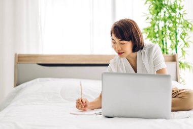 Genç gülümseyen mutlu Asyalı kadın evdeki yatak odasında dizüstü bilgisayar kullanarak rahatlıyor. Ev konseptinden çalışıp klavyede yazan genç yaratıcı kız.