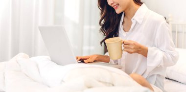 Genç gülümseyen mutlu Asyalı kadın dizüstü bilgisayar kullanarak rahatlıyor ve evdeki yatak odasında kahve içiyor. Genç yaratıcı kız klavyede çalışıyor ve daktilo kullanıyor. Ev konseptinden çalışıyor.
