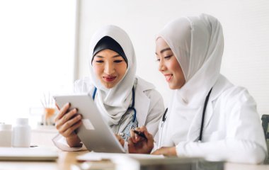 Hastane masasında pano ve tablet bilgisayarla çalışan iki Asyalı Müslüman kadın doktor. Sağlık ve ilaç servisi.
