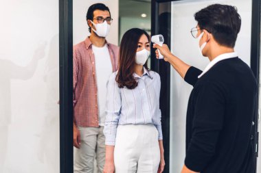 Bir grup profesyonel Asya işi ve ofiste koruyucu maske takarak koronavirüs için karantinada çalışmadan önce vücut ısısını ölçmek için kızılötesi termometre kullanılıyor.