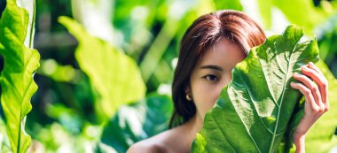 Gülümseyen güzel Asyalı güzel bir kadının portresi. Temiz, taze, sağlıklı bir beyaz ten. Bulanık yeşil arka planda yeşil yaprağın arkasında rahatlayın.