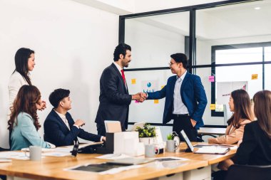 Zarif takım elbiseli iki Asyalı iş ortağının modern ofisteki gündelik işler grubunun önünde el sıkışmasını hayal edin. Ortaklık onayı ve teşekkür jest konsepti.