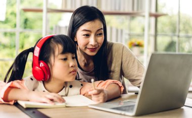 Anne ve Asyalı küçük kız öğreniyor ve dizüstü bilgisayara bakıyor ev ödevi yapıyor çevrimiçi eğitim e-öğrenme sistemiyle bilgi öğreniyor. Evde öğretmenle çocuk video konferansı