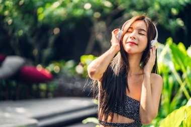 Gülümseyen, güzel Asyalı güzel bir kadının kulaklıklarıyla durup müzik dinlemesi rahatla ve doğa ile temiz havanın tadını çıkar.